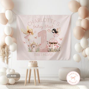 May include: A pink birthday banner with the text "CHARLOTTE'S fairy first BIRTHDAY" and illustrations of fairies, flowers, and a mushroom. Balloons in white and blush pink are on either side of the banner. A small cake sits on a white table.