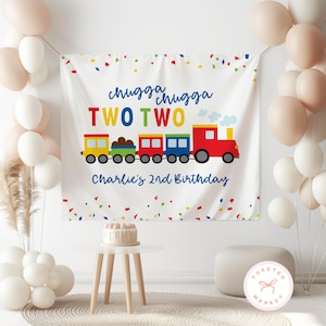 May include: A white banner with a colorful train illustration and the text "chugga chugga TWO TWO Charlie's 2nd Birthday". The banner is decorated with confetti and surrounded by balloons. A small cake sits on a white table.