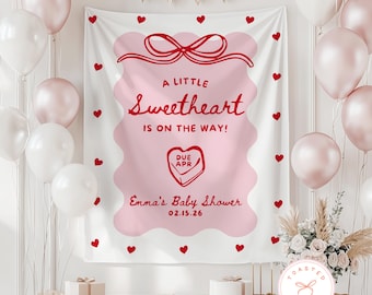 May include: A baby shower backdrop with the text "A Little Sweetheart Is On The Way!" in red script. The backdrop is pink with a scalloped edge and red heart accents. White and pink balloons and a floral arrangement complete the scene.