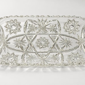 May include: A clear rectangular glass tray featuring intricate cut patterns including multiple starburst motifs, geometric triangular shapes, and a scalloped edge. The surface displays a complex arrangement of faceted glass elements that reflect light across the entire piece.