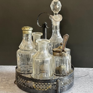 May include: A vintage condiment set featuring a circular dark metal stand with a central handle. The set includes four clear glass containers: a shaker with a metal lid, two small bottles, and a lidded jar with a metal spoon inside.