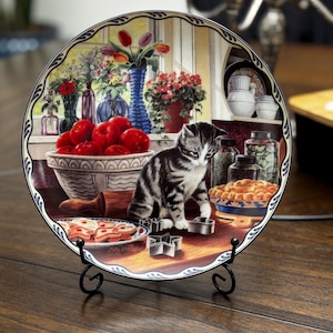 May include: Decorative plate featuring a kitten in a kitchen scene with apples, flowers, and baked goods. The plate has a scalloped edge and is displayed on a black metal stand. The artwork includes a bowl of red apples and a pie.