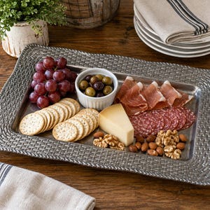 May include: A rectangular metal serving tray with a textured, woven-style border. The tray holds a charcuterie arrangement featuring red grapes, a small white bowl of green and black olives, crackers, sliced prosciutto, salami, cheese, and nuts.