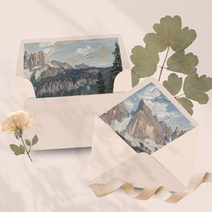 May include: Two cream envelopes, each containing a landscape painting of mountain ranges under a blue sky with white clouds. A dried flower and green leaves are arranged near the envelopes, with a cream ribbon. The paintings are in an oil painting style.