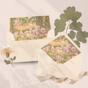 May include: Two cream-colored envelopes containing cards with a printed landscape illustration of pink and white flowers in a garden. A single dried flower and a sprig of green leaves are placed nearby, accompanied by a light beige ribbon.