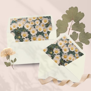 May include: Two cream-colored envelopes with daisy floral cards inside, arranged on a light pink surface. A dried flower and green leaves are placed near the envelopes. A beige ribbon is draped across the bottom.