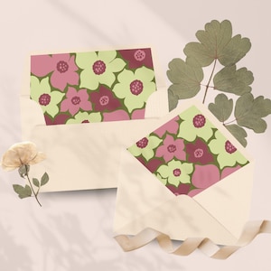 Puede incluir: Dos sobres de color crema que contienen tarjetas con un estampado floral en tonos verde claro, rosa y granate oscuro. Las tarjetas presentan flores estilizadas de cinco pétalos con centros circulares. Incluye una hoja verde seca y una flor pequeña.