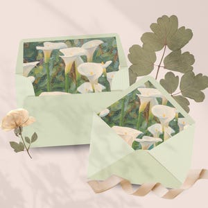 May include: Two pale green envelopes, each displaying a painting of white calla lilies. A dried flower and a sprig of leaves are positioned near the envelopes. A beige ribbon is draped across the bottom.