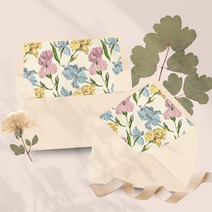 Peut inclure: Deux enveloppes crème avec des motifs floraux d'iris roses, bleus et jaunes. L'une des enveloppes est ouverte, révélant le motif floral. Une fleur séchée et des feuilles vertes sont disposées à proximité, avec un ruban crème.