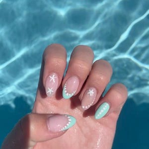 May include: A hand with almond-shaped nails, each painted with a unique design. The nails feature a light pink base with a white dotted border and a light blue tip. Some nails have white star accents.