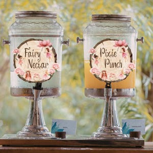 May include: Two glass drinks dispensers with silver taps and lids. Each has a wooden-effect label with the text "Fairy Nectar" and "Pixie Punch", surrounded by pink flowers and fairy illustrations. The dispensers are on a wooden tray.