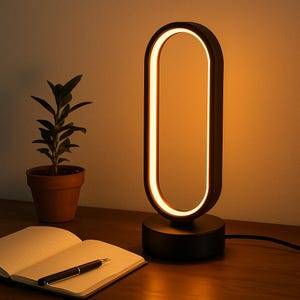 Könnte beinhalten: Eine moderne, schwarze, ovale Tischlampe mit warmem, leuchtendem Licht. Die Lampe steht auf einem runden Sockel und ist an eine Steckdose angeschlossen. Eine kleine Topfpflanze, ein offenes Notizbuch und ein Stift befinden sich ebenfalls auf der Holzoberfläche.