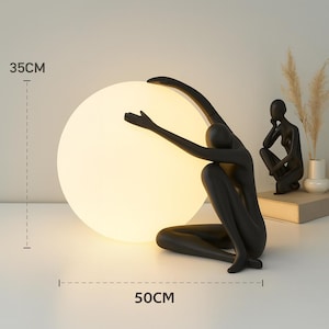 Peut inclure: Une sculpture noire d'une personne agenouillée et tendant les bras vers une grande lumière sphérique et brillante. La lampe mesure 50 cm de large et 35 cm de haut. Une petite sculpture noire d'une personne assise est à l'arrière-plan.