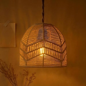 May include: A woven pendant light fixture with a natural, beige color. The light has a dome shape with a woven pattern and a single light bulb visible inside. The cord is a twisted rope style.