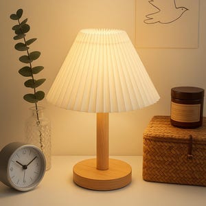 May include: A cream-colored pleated lampshade sits atop a wooden base, casting a warm glow. A small analog clock, a woven basket, and a jar with a label are also on the table. A drawing of a bird is on the wall.