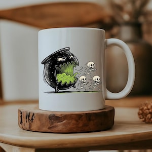 May include: A white ceramic mug featuring a graphic illustration of a black, sharp-toothed cauldron with a wide, open mouth emitting a bright green liquid. Three small, white skeletons are depicted running away from the cauldron on the mug surface.