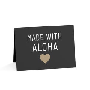 May include: A rectangular folded greeting card with a dark charcoal gray background. The text "MADE WITH ALOHA" is printed in a clean, white, sans-serif font. Below the text, there is a solid, light tan-colored heart icon centered on the card.