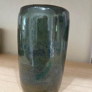Op de afbeelding: Een handgemaakte keramische beker met een glanzend, gevlekt glazuur in groen- en bruintinten. De cilindrische beker heeft een enigszins onregelmatige vorm, wat bijdraagt aan zijn rustieke charme. De binnenkant is donkerder groen en de buitenkant heeft een gradiënteffect.