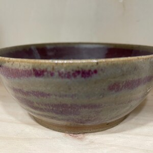 May include: A handcrafted ceramic bowl with a wide, shallow form. The bowl has a muted colour palette with a light beige exterior and a deep burgundy interior. Horizontal bands of burgundy accent the exterior.