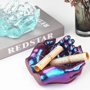 May include: A decorative iridescent purple and blue hand-shaped dish with two rolled scrolls inside. The dish is made of resin and has a smooth, glossy finish.