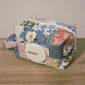 May include: A patchwork fabric wipe case with a white lid that says "HUGGIES". The case is rectangular with a zipper closure and a small handle. The fabric is a mix of floral and patterned designs in blue, pink, and white.
