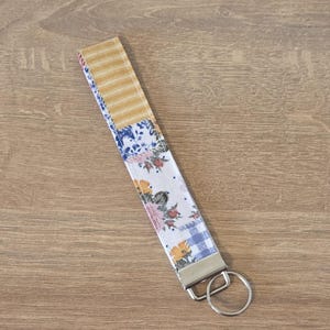 May include: A fabric wristlet key fob with a patchwork design. The fob features sections of yellow stripes, blue floral patterns, and a blue and white gingham check. A silver-colored metal ring is attached for keys.