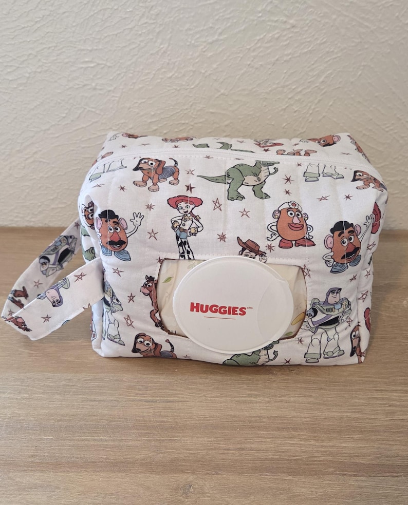 May include: A white fabric wipe dispenser with a pattern of cartoon characters. The dispenser has a handle and a circular opening with the word "HUGGIES" printed on it. The design includes characters like Woody and Buzz Lightyear.