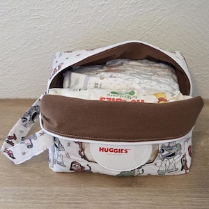 May include: A white and brown nappy bag with a handle, decorated with a cartoon character pattern. The bag is open, showing nappies and a container of Huggies wipes. The interior is brown, and the bag is on a light wood surface.