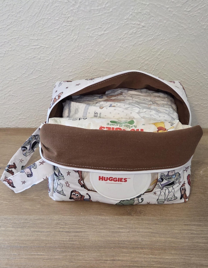 May include: A white and brown nappy bag with a handle, decorated with a cartoon character pattern. The bag is open, showing nappies and a container of Huggies wipes. The interior is brown, and the bag is on a light wood surface.