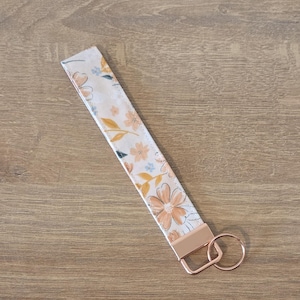 May include: A floral patterned wristlet key fob with a rose gold metal clasp and key ring. The fabric features peach and blue flowers on a cream background. The key fob is laying on a wooden surface.