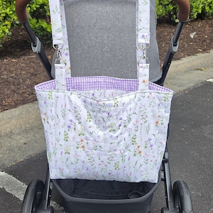 May include: A white and lavender floral print stroller organizer bag with a purple gingham lining. The bag is attached to a gray stroller with brown leather handles. The bag has straps with silver metal clips.
