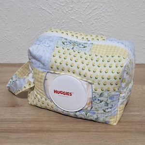 May include: A quilted fabric pouch featuring a patchwork design of yellow floral, blue floral, and yellow gingham patterns. The pouch includes a white plastic Huggies brand wipe dispenser lid centered on the front and a side carrying strap.