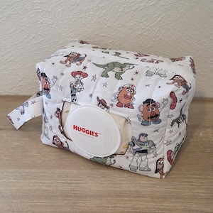 May include: A white, rectangular baby wipes container with a zipper closure and a loop handle. The container is covered in a pattern of cartoon characters and stars. A white Huggies wipes dispenser is visible.