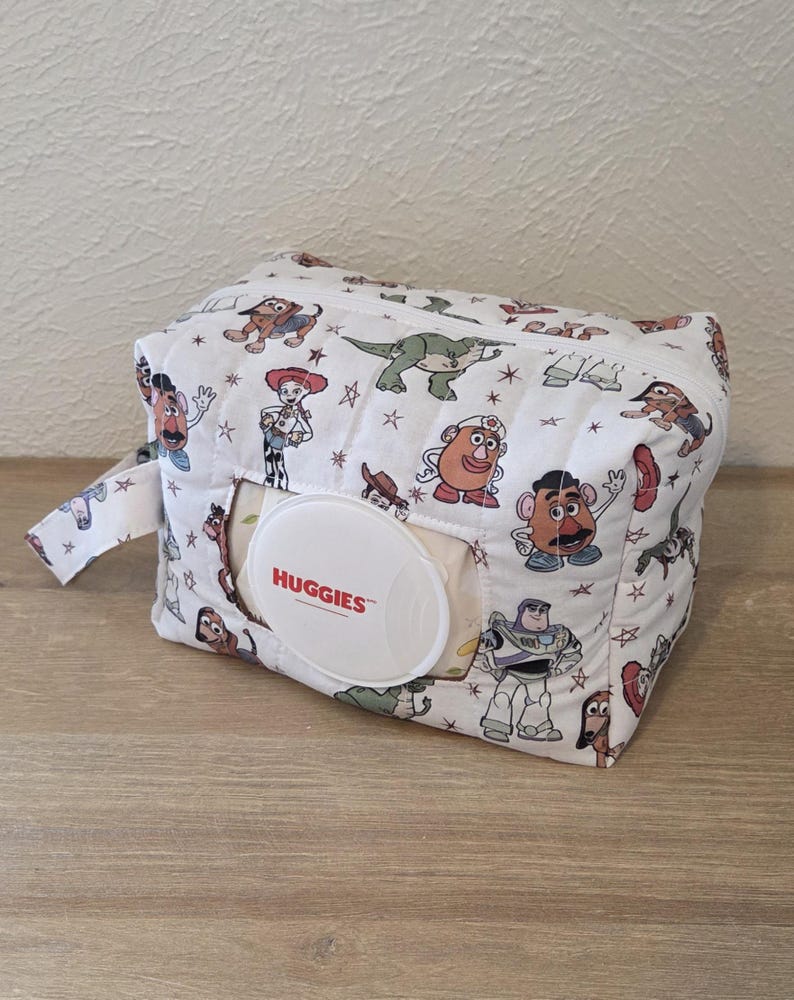 May include: A white baby wipes container with a zip and loop handle. The container is patterned with cartoon characters and stars. A white Huggies wipes dispenser is visible, ideal for baby care.
