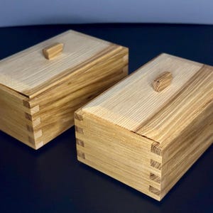 May include: Two rectangular wooden boxes with lids. The boxes are light-colored with visible wood grain and dovetail joints. Each box has a small, rectangular wooden knob on the lid. The boxes are on a dark surface.