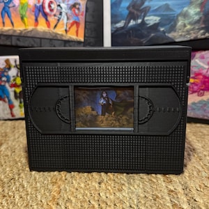 May include: A black, rectangular, 3D-printed storage box designed to resemble a vintage VHS cassette tape. The front features a central rectangular window displaying a small, colorful illustration of a character standing in a rocky landscape.