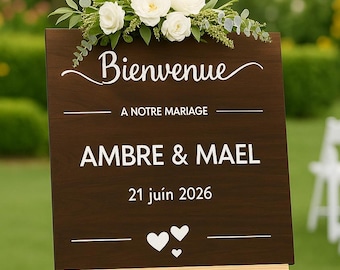 May include: A wooden wedding welcome sign on an easel, with the words "Bienvenue" and "AMBRE & MAEL 21 juin 2026". The sign is decorated with white roses and greenery. The sign is displayed outdoors.