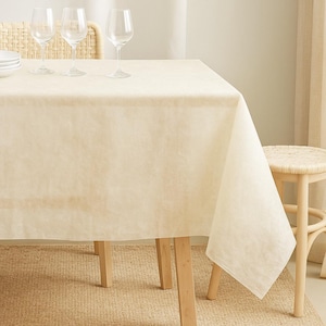 May include: A cream-coloured rectangular tablecloth on a wooden table. Three wine glasses and a stack of white plates are visible. A woven chair and stool are in the background, creating a neutral-toned dining setting.