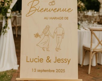 Può includere: Un cartello di nozze in legno su un cavalletto. Il cartello recita "Bienvenue" e "Au Mariage De Lucie & Jessy 13 settembre 2025" in lettere dorate. È presente anche un disegno a tratto di una coppia che si tiene per mano.