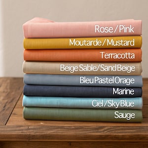 May include: A stack of folded fabric in various colors, including pink, mustard, terracotta, sand beige, pastel blue, navy, sky blue, and sage. Each fabric is labeled with its color name. The fabrics are stacked on a wooden surface.