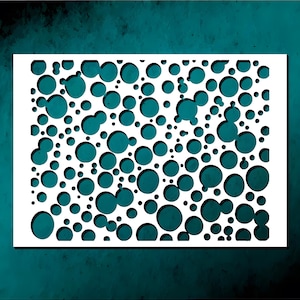 May include: A white stencil with a pattern of varying sized circles cut out. The circles are scattered across the rectangular shape, creating a random, abstract design. The background is a teal color.