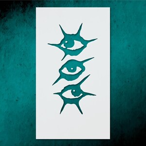 May include: A white rectangular stencil featuring three stylized eye designs cut out in teal. Each eye is surrounded by spiky, star-like shapes. The background is a textured teal color.