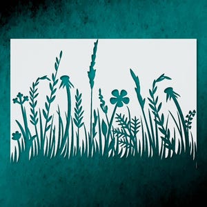 May include: A white paper stencil featuring a detailed silhouette of a meadow scene. The design includes various types of grass, flowers, and seed heads, all cut out to create a negative space effect. The background is a teal color.