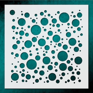 May include: A white stencil with a pattern of varying sized circles cut out. The background is a teal color. The circles are of different sizes and are scattered across the stencil. The stencil is square in shape.