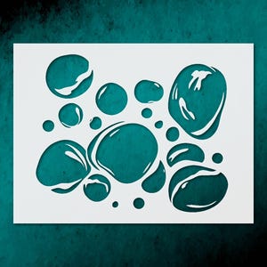May include: White stencil featuring a pattern of various sized water droplets. The droplets are cut out, revealing a teal background. The stencil is rectangular and suitable for crafts and DIY projects.
