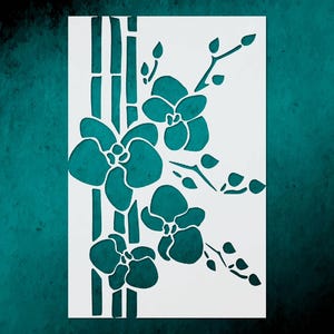 May include: A white stencil featuring a teal orchid design with bamboo stalks. The orchid flowers and leaves are cut out, creating a negative space effect. The design is set against a teal background.