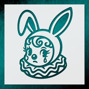 May include: A teal-colored stencil of a sad bunny face with a clown collar. The bunny has large eyes, long eyelashes, and a teardrop. The design is cut out of a white square.