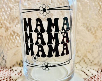 May include: A clear glass can-shaped drinking glass with the word "MAMA" in black, stacked three times, inside a black rectangular frame. Small daisy flower graphics are at each corner of the frame. The glass sits on a white saucer.