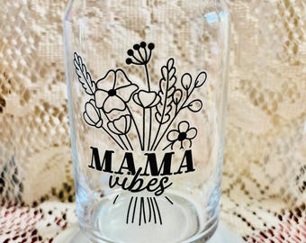 May include: A clear glass can-shaped drinking glass with a white base. The glass features a black floral design and the words "MAMA vibes" in a stylish font. The glass is empty and sits on a white, scalloped-edge plate.