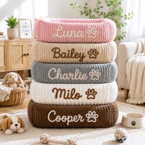 May include: A stack of five rectangular pet beds made of ribbed corduroy fabric in pink, beige, gray, white, and brown. Each bed features embroidered names: Luna, Bailey, Charlie, Milo, and Cooper, accompanied by a small paw print icon.